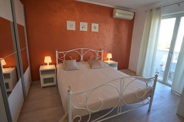 A bedroom with a bed, two bedside tables with lamps, a wall-mounted air conditioner, and a sliding glass door to the right.