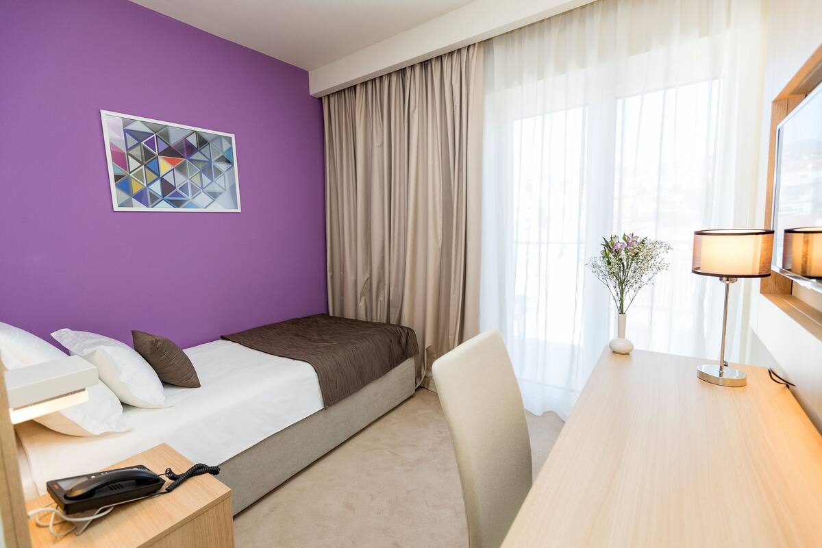 Een hotelkamer met een bed, een bureau, een lamp, een telefoon, een vaas met bloemen en een schilderij aan de muur.