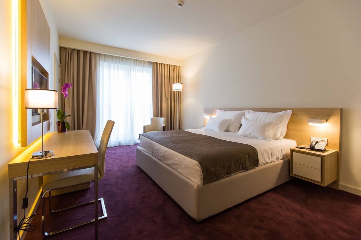 Hotelkamer met een bed, nachtkastje, lamp, bureau, stoel en een groot raam met gordijnen.