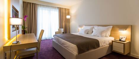 A hotel room with a bed, bedside table, lamp, desk, chair, and a large window with curtains.