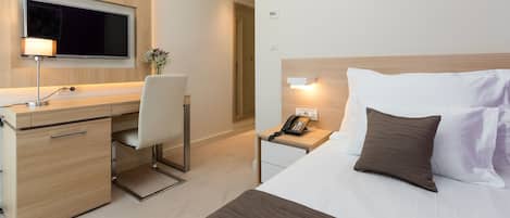 A hotel room with a desk, chair, bed, bedside table, lamp, and a flat-screen TV mounted on the wall.