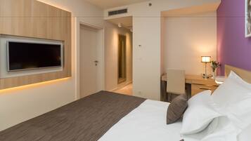 A hotel room with a bed, a desk, a chair, a TV mounted on the wall, and a lamp.