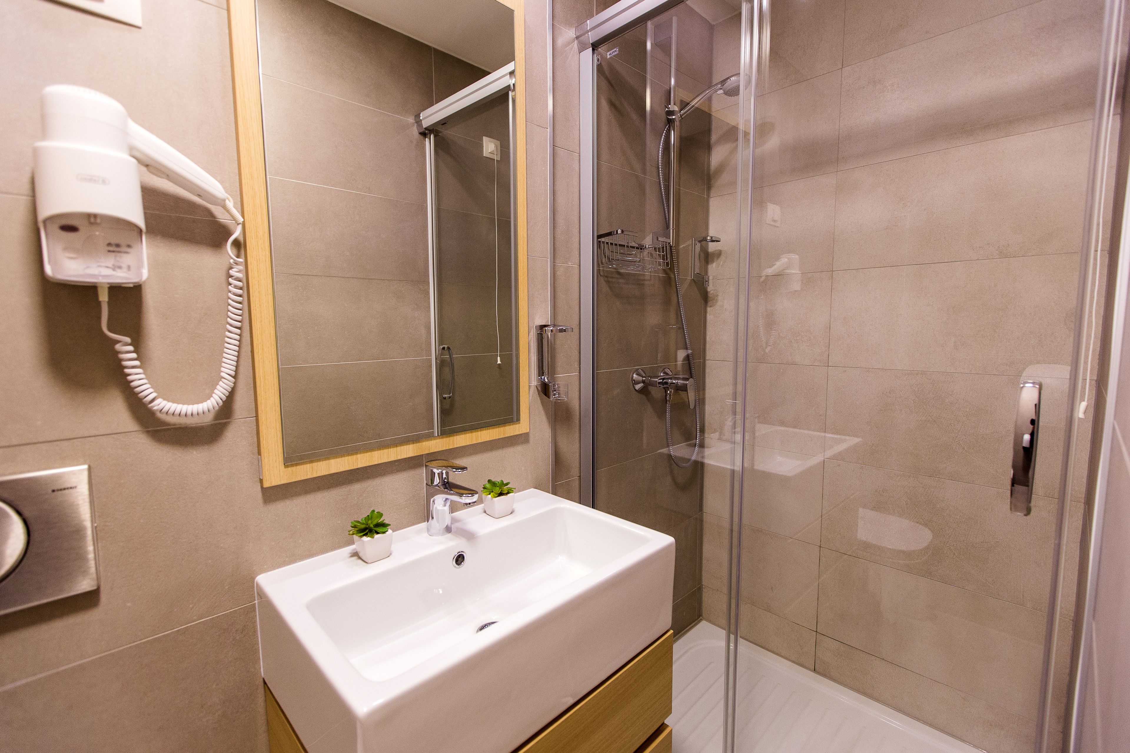 Un baño moderno con un lavamanos blanco, mampara de ducha de vidrio y un secador de pelo montado en la pared.