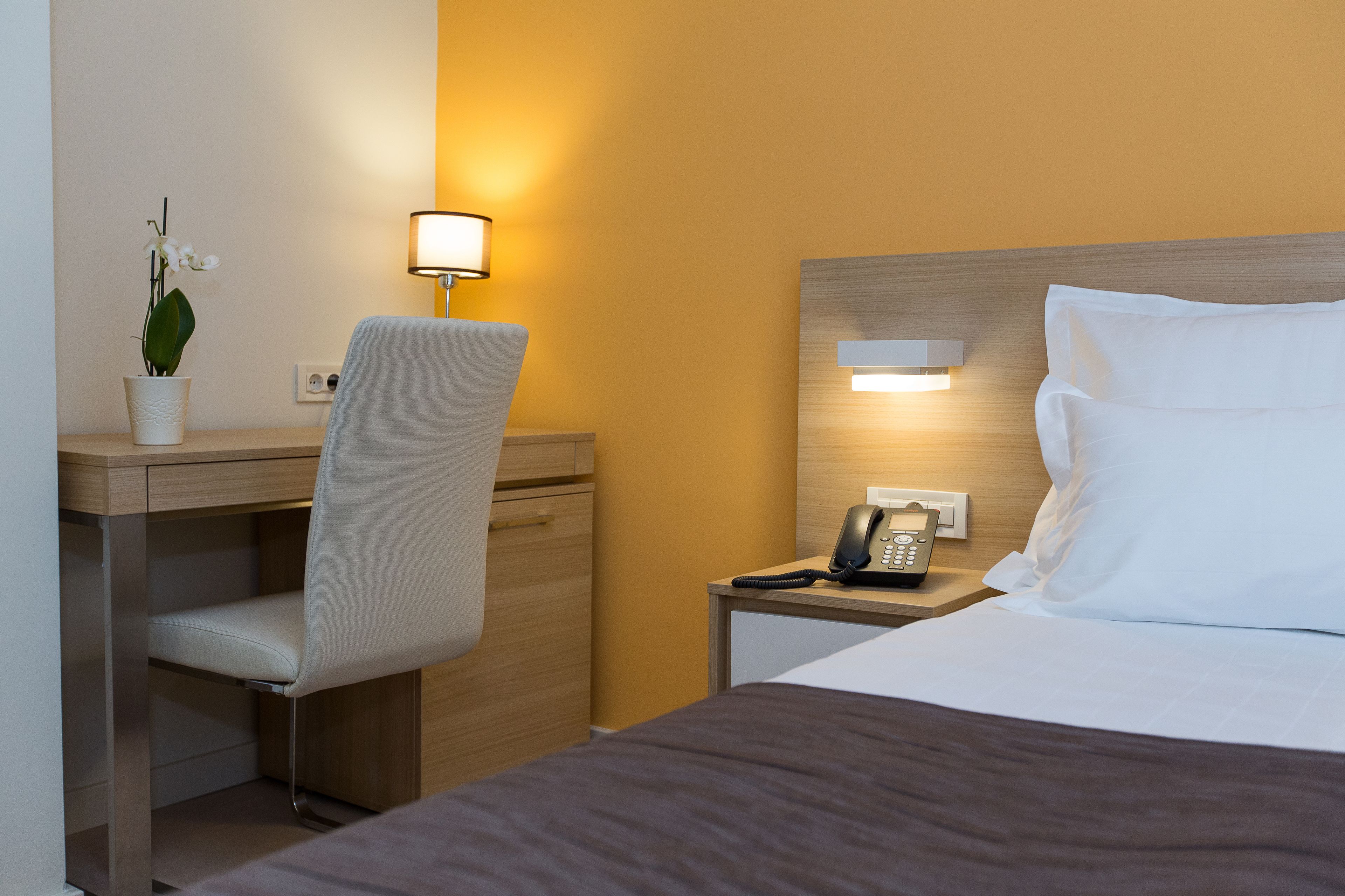 Een hotelkamer met een bed, een bureau met een stoel, een nachtkastje met een telefoon en een wandlamp.