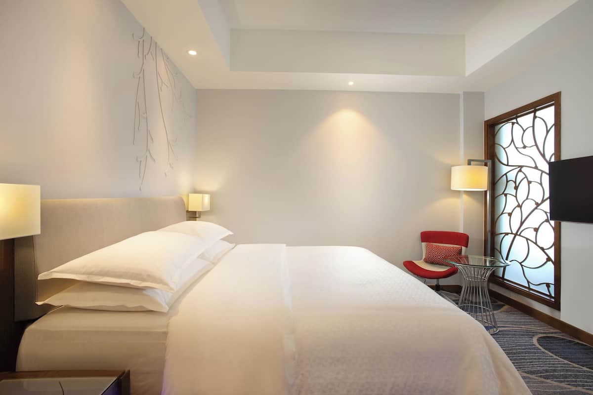 A hotel room with a large bed, two bedside lamps, a small table, a red chair, and a decorative screen.