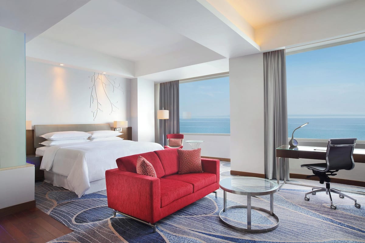 A hotel room with a bed, a red sofa, a desk with a chair, a round table, and a view of the sea.