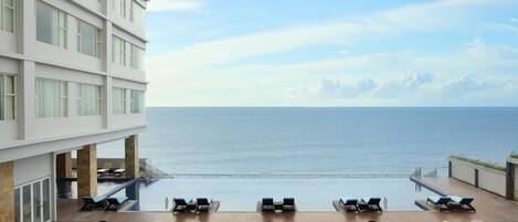 A modern hotel with a rooftop infinity pool overlooking the sea, lounge chairs, and a dining area.