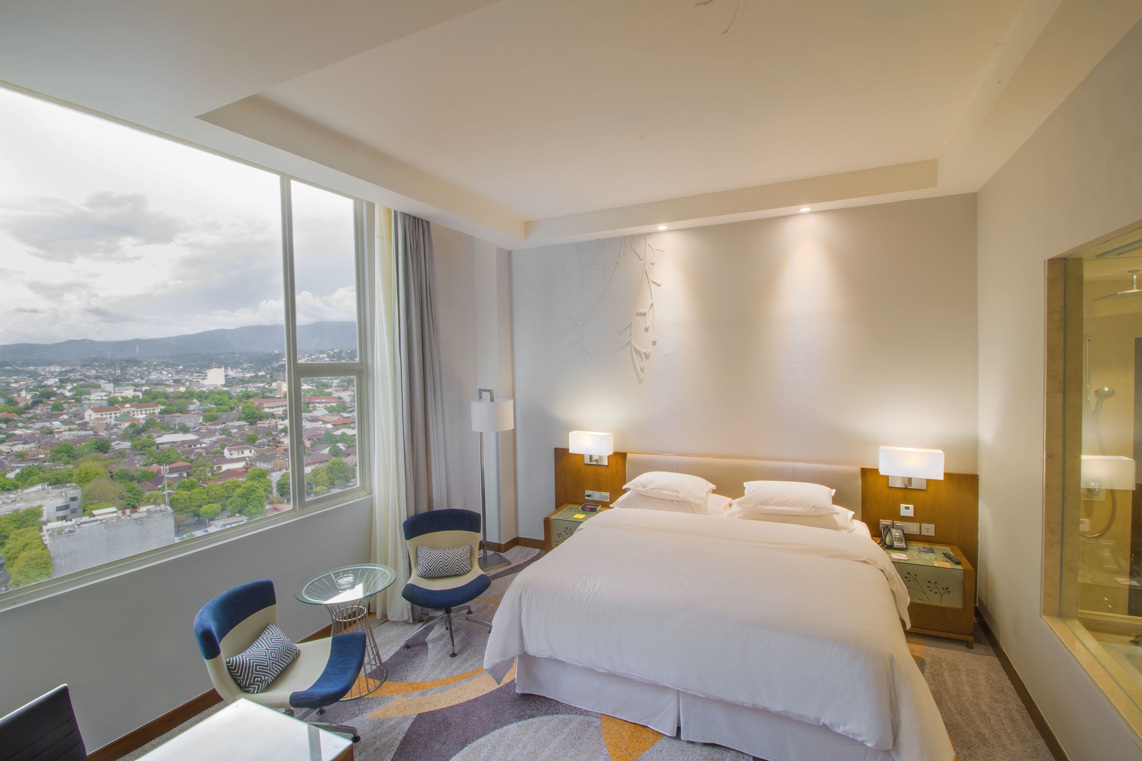 A hotel room with a large bed, two bedside tables, a chair, a small table, and a view of a cityscape through a large window.