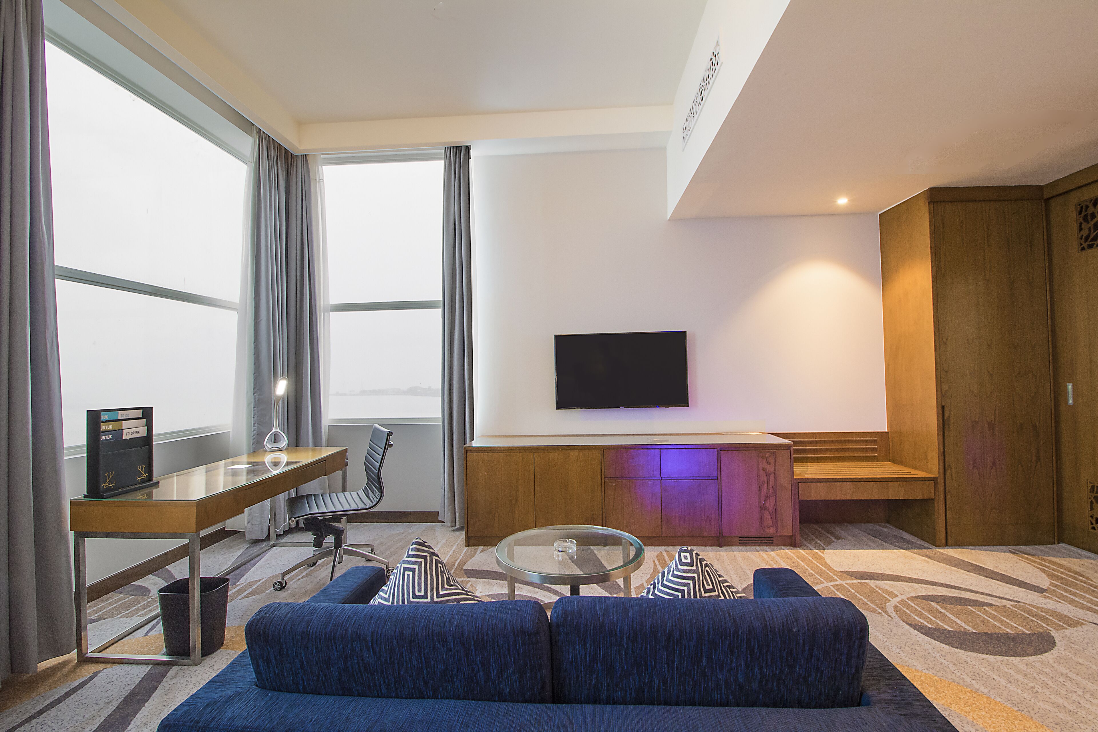 A modern hotel room with a large window, a flat-screen TV, a sofa, a desk, and a wooden cabinet.