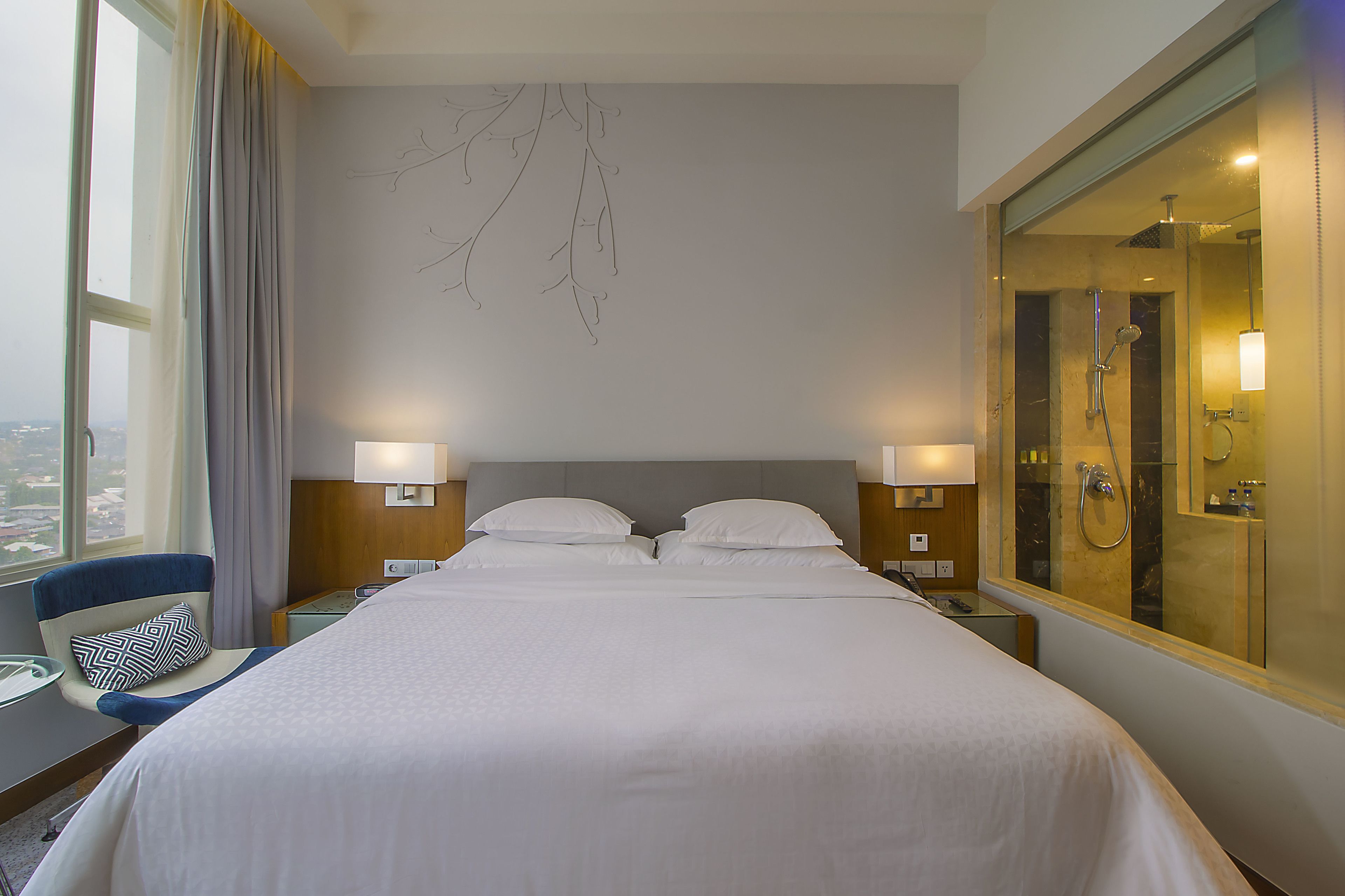 Habitació d'hotel amb un llit gran, dues taules de nit, una cadira, una finestra amb cortines i un bany amb plat de dutxa de vidre.