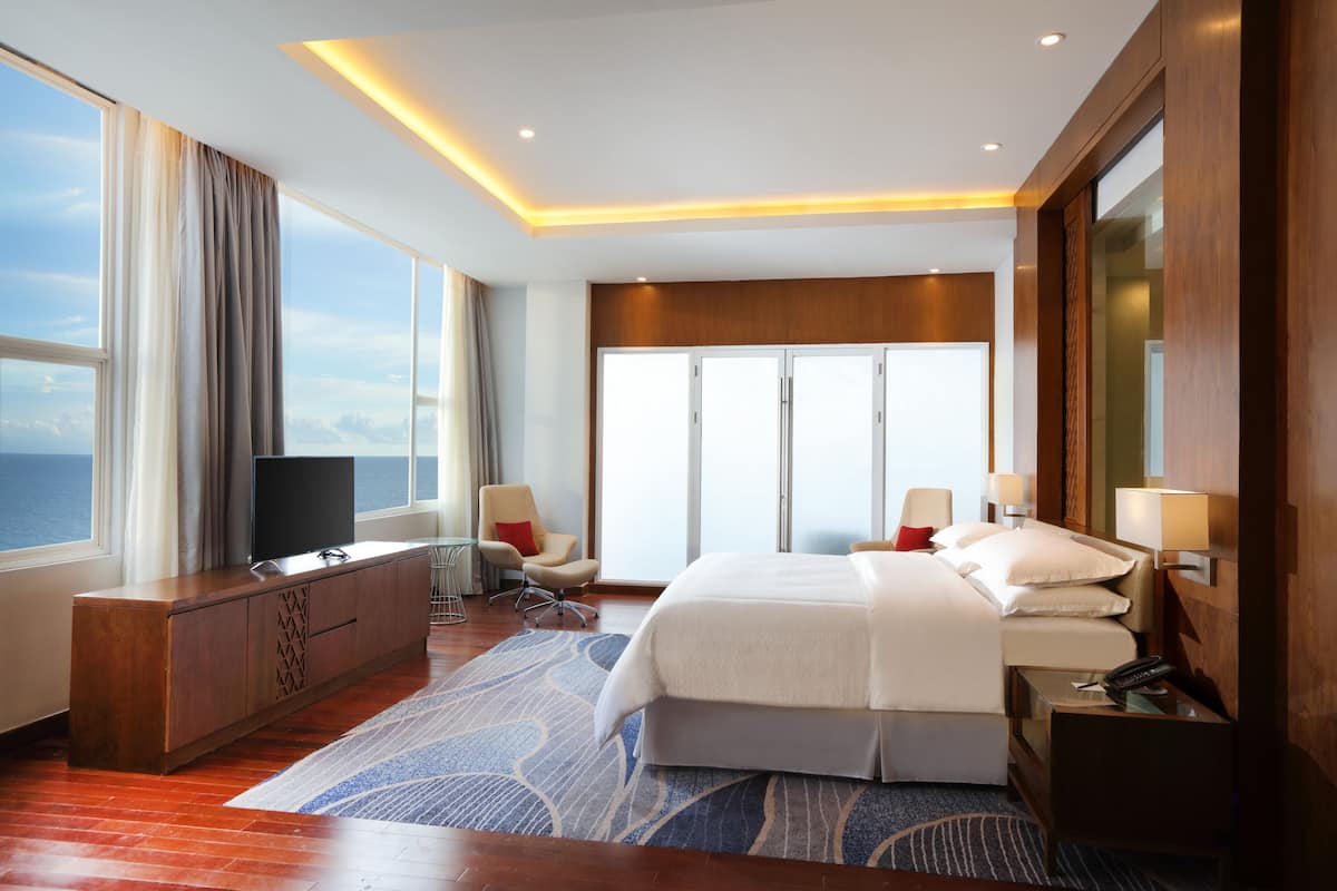 A hotel room with a large bed, a TV on a wooden cabinet, a desk with a lamp, and a view of the ocean through the windows.