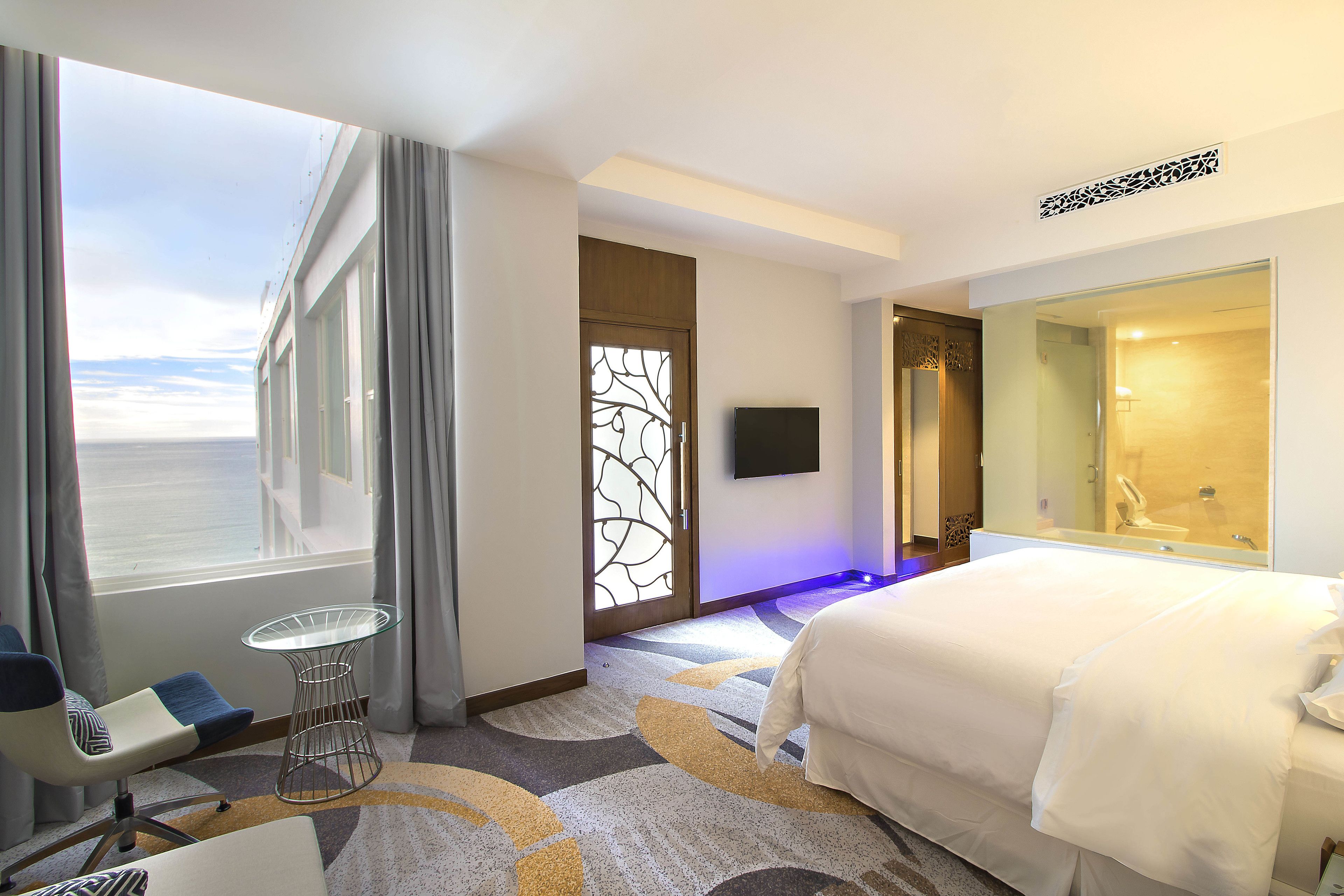 Una camera d'albergo con un letto grande, un tavolino in vetro, una sedia, una televisione e vista sul mare.