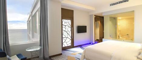 Habitación de hotel con una cama grande, una mesa de vidrio, una silla, televisión y vista al mar.