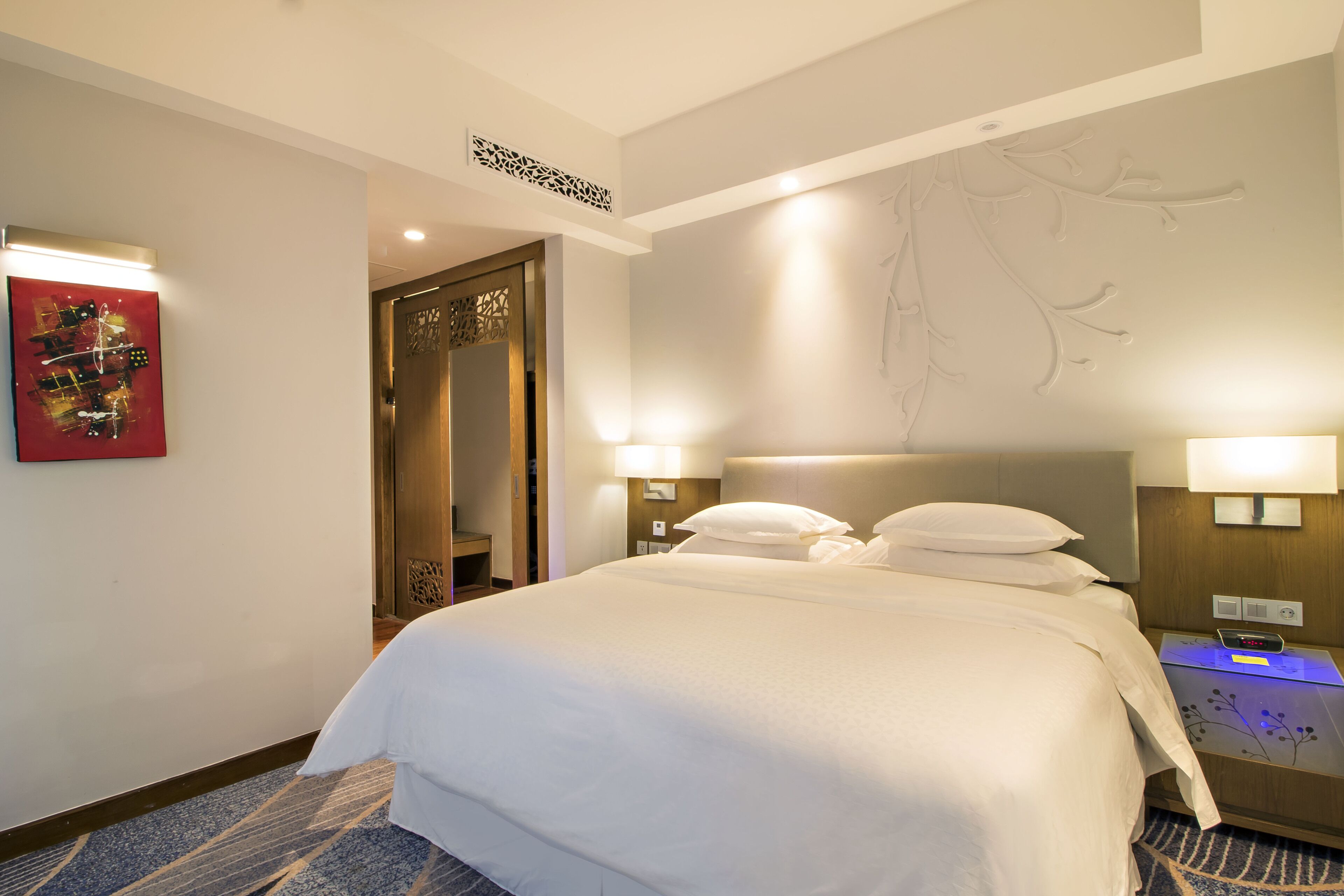 A hotel room with a large bed, two bedside tables with lamps, a red abstract painting, and a decorative ceiling design.