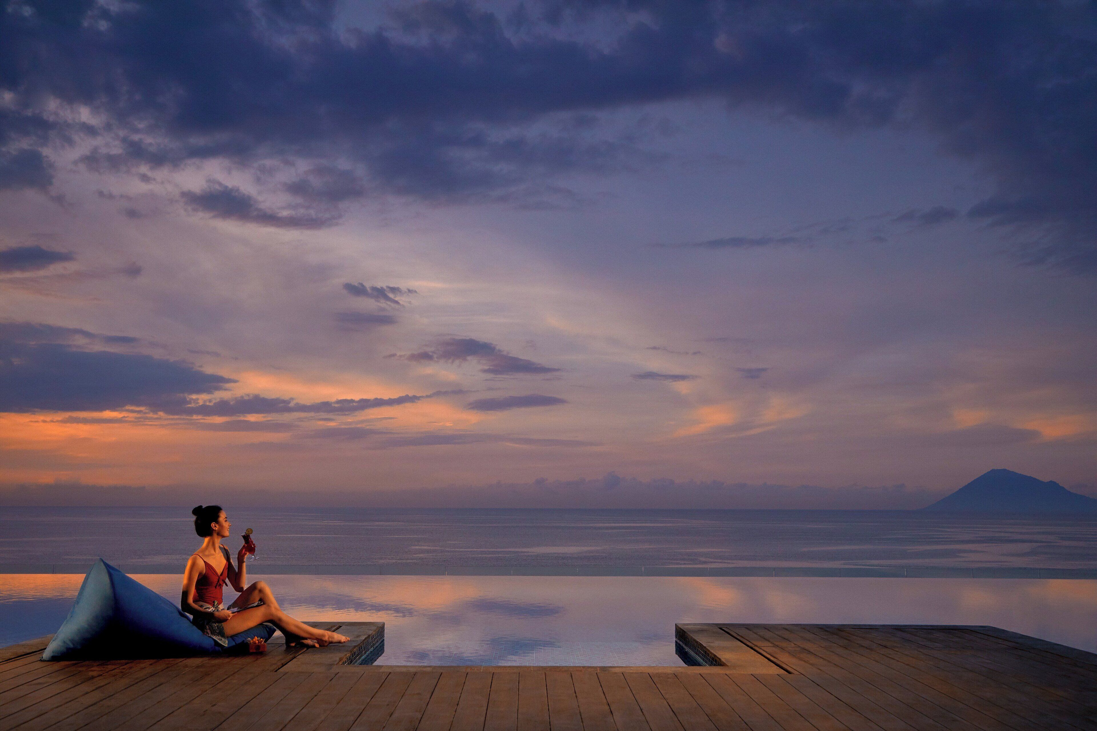 A person sitting on a wooden deck by an infinity pool with a mountain in the background.