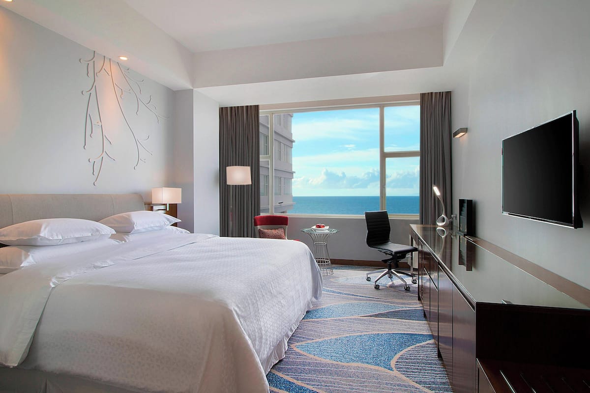 A hotel room with a large bed, a desk with a chair, a TV, and a view of the ocean.