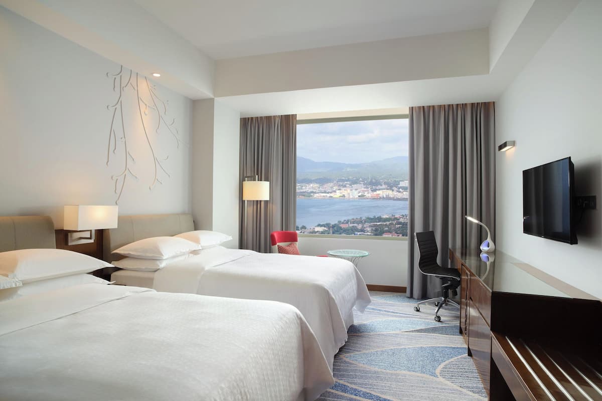 A hotel room with two beds, a TV, a desk, and a view of a city and mountains.