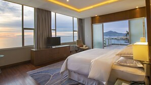 Habitación de hotel con una cama grande, televisión, escritorio con silla y vista al mar y a las montañas.