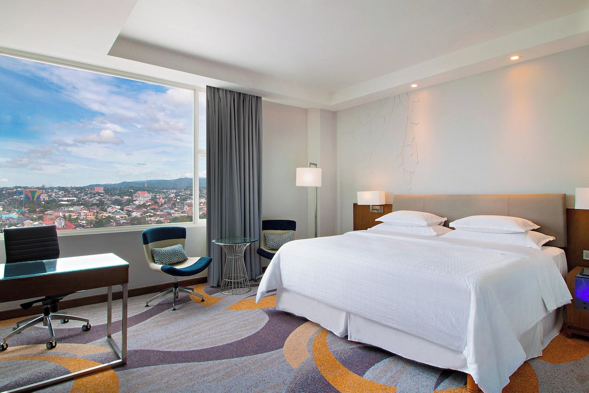 A hotel room with a large bed, a desk, a chair, a small table, and a view of a cityscape through a window.