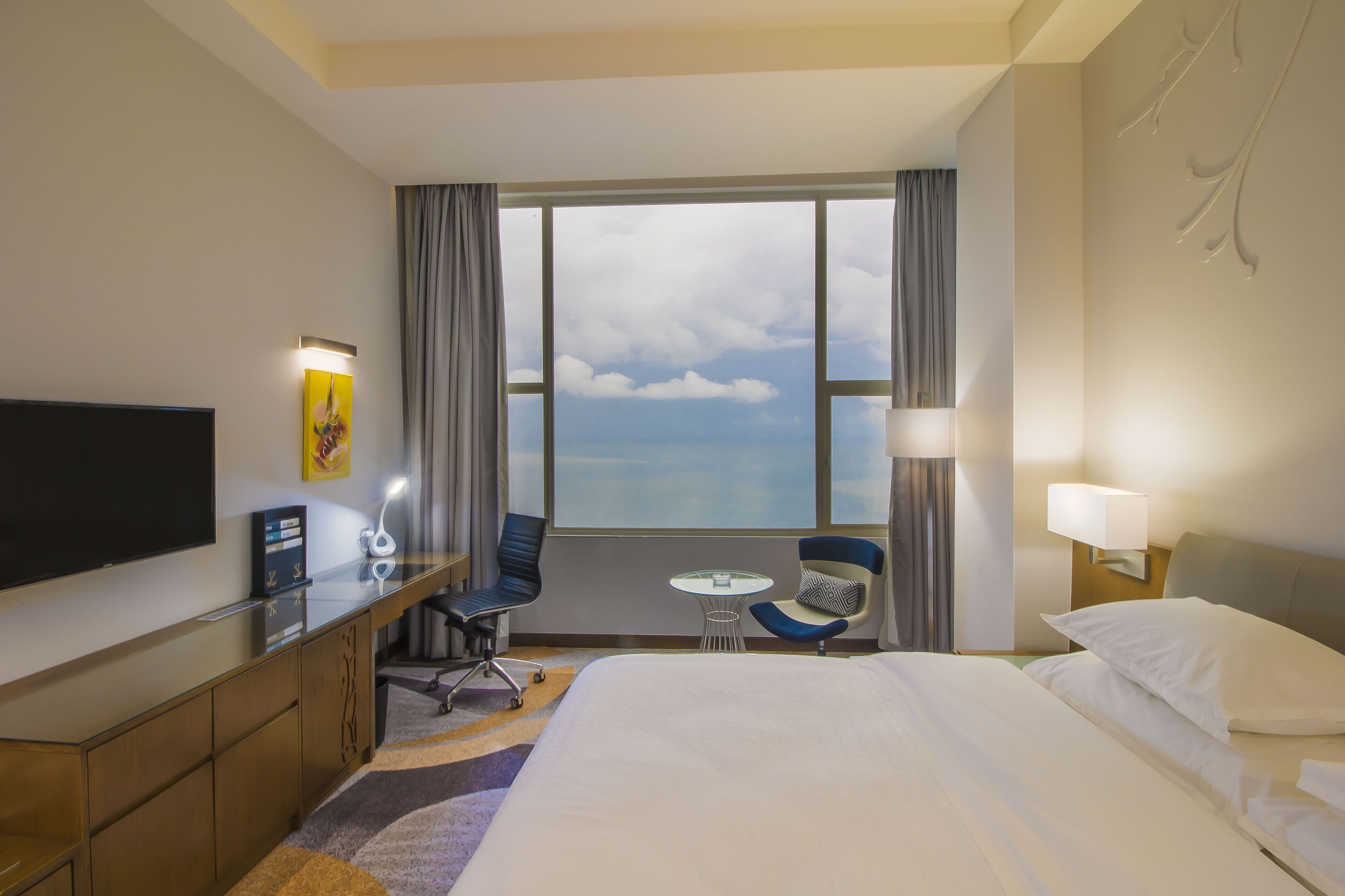 A hotel room with a bed, a desk with a flat-screen TV, a small table, two chairs, a window with a view of the sky, and a wall-mounted lamp.