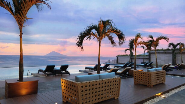 Terraza con sillones de descanso y una jardinera de madera, con vista a una masa de agua con palmeras y un atardecer.