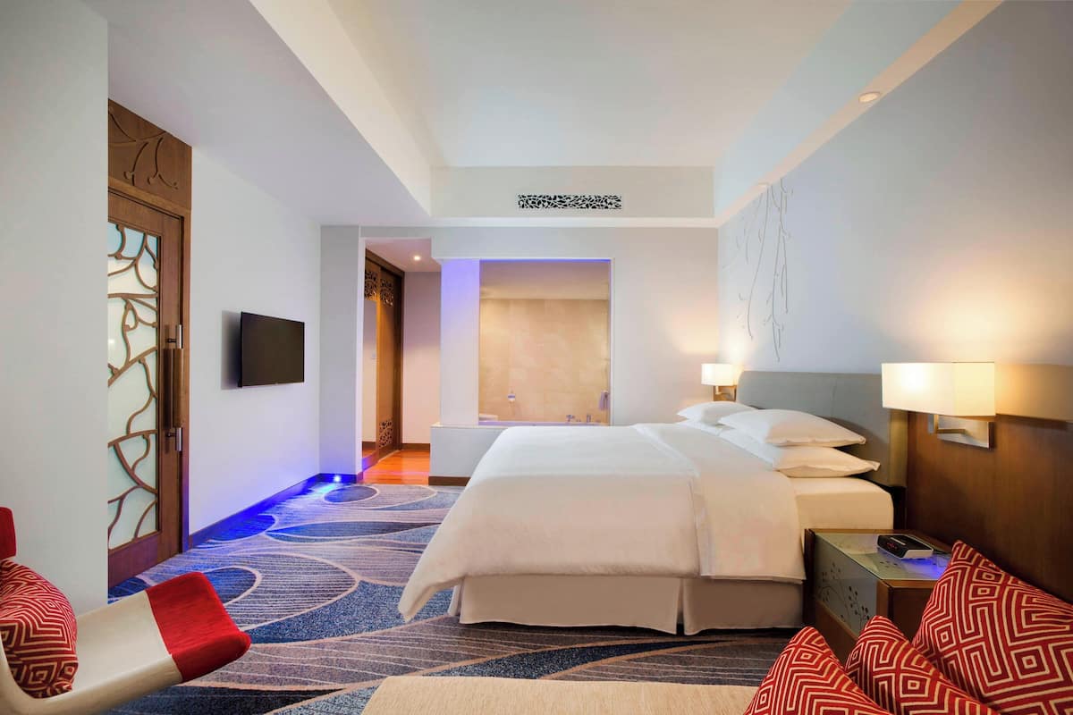 A hotel room with a large bed, a TV mounted on the wall, a seating area with a red and white chair, and a patterned carpet.