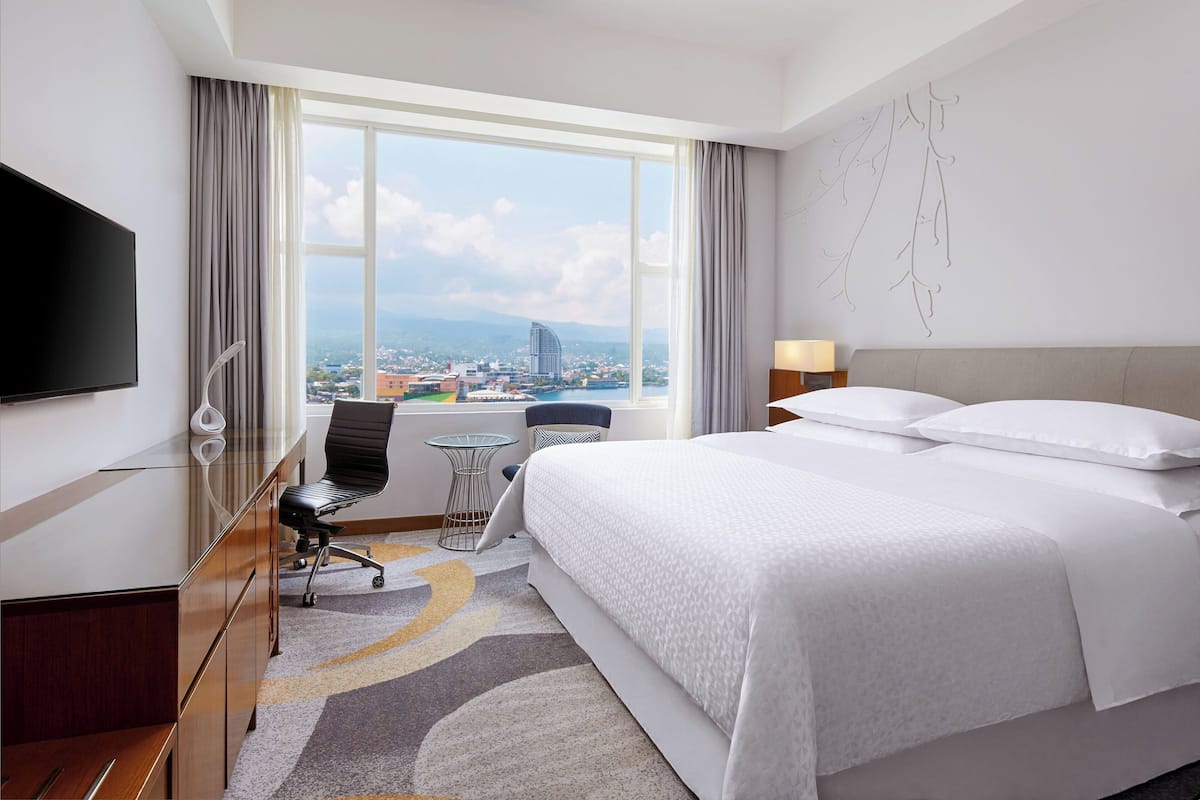 A hotel room with a large bed, a desk with a chair, a TV, and a view of a cityscape through a large window.