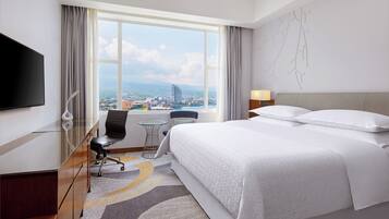 A hotel room with a large bed, a desk with a chair, a TV, and a view of a cityscape through a large window.