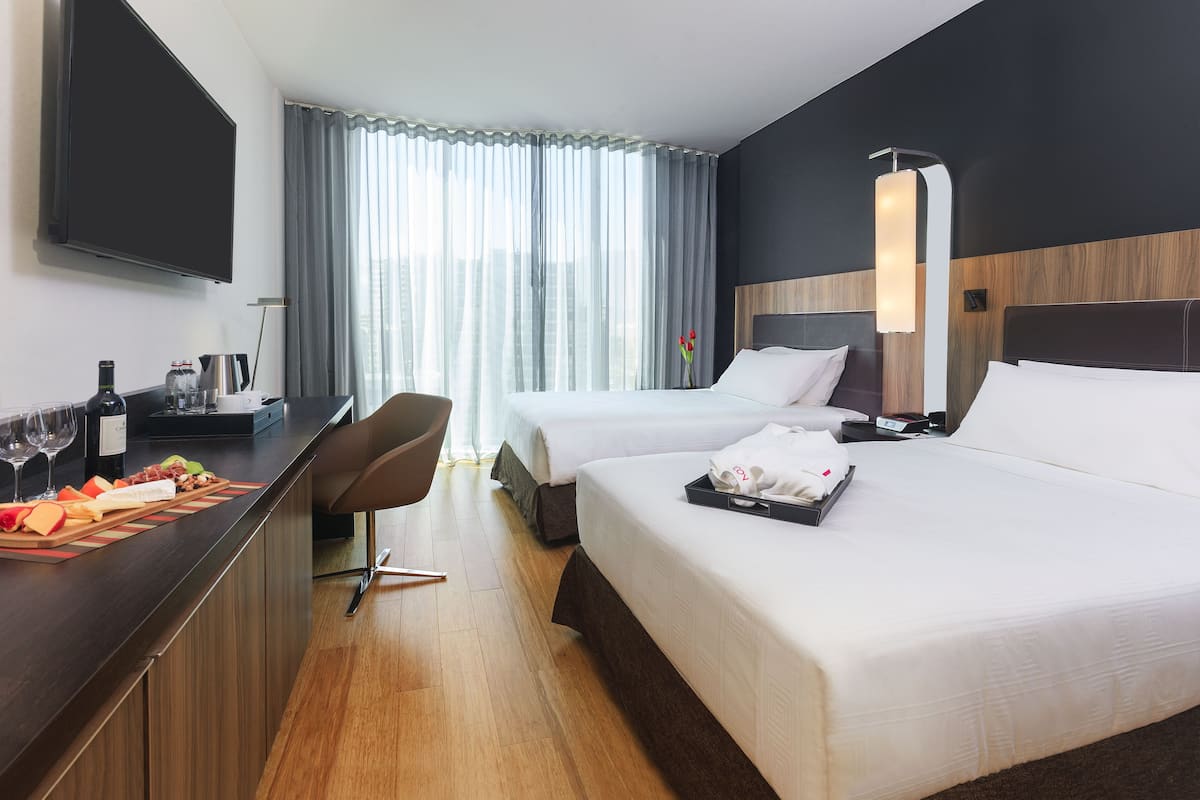A hotel room with two beds, a desk with a flat-screen TV, a minibar, and a large window with curtains.