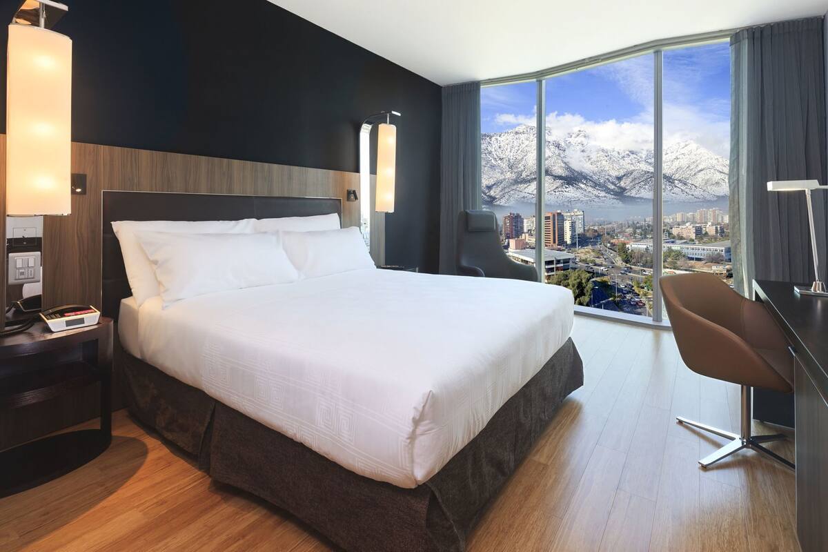 A hotel room with a large bed, a desk, and a view of a cityscape through a large window.
