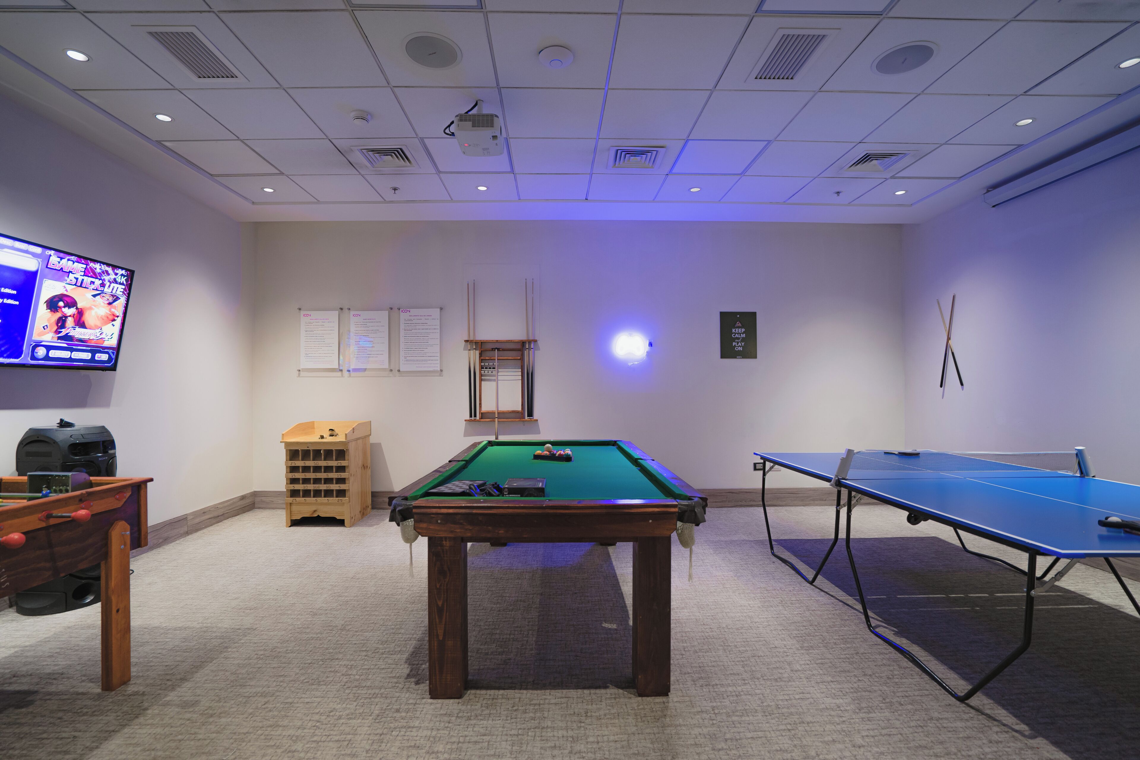 A room with a pool table, a foosball table, a billiards table, and a projector screen displaying a video game.
