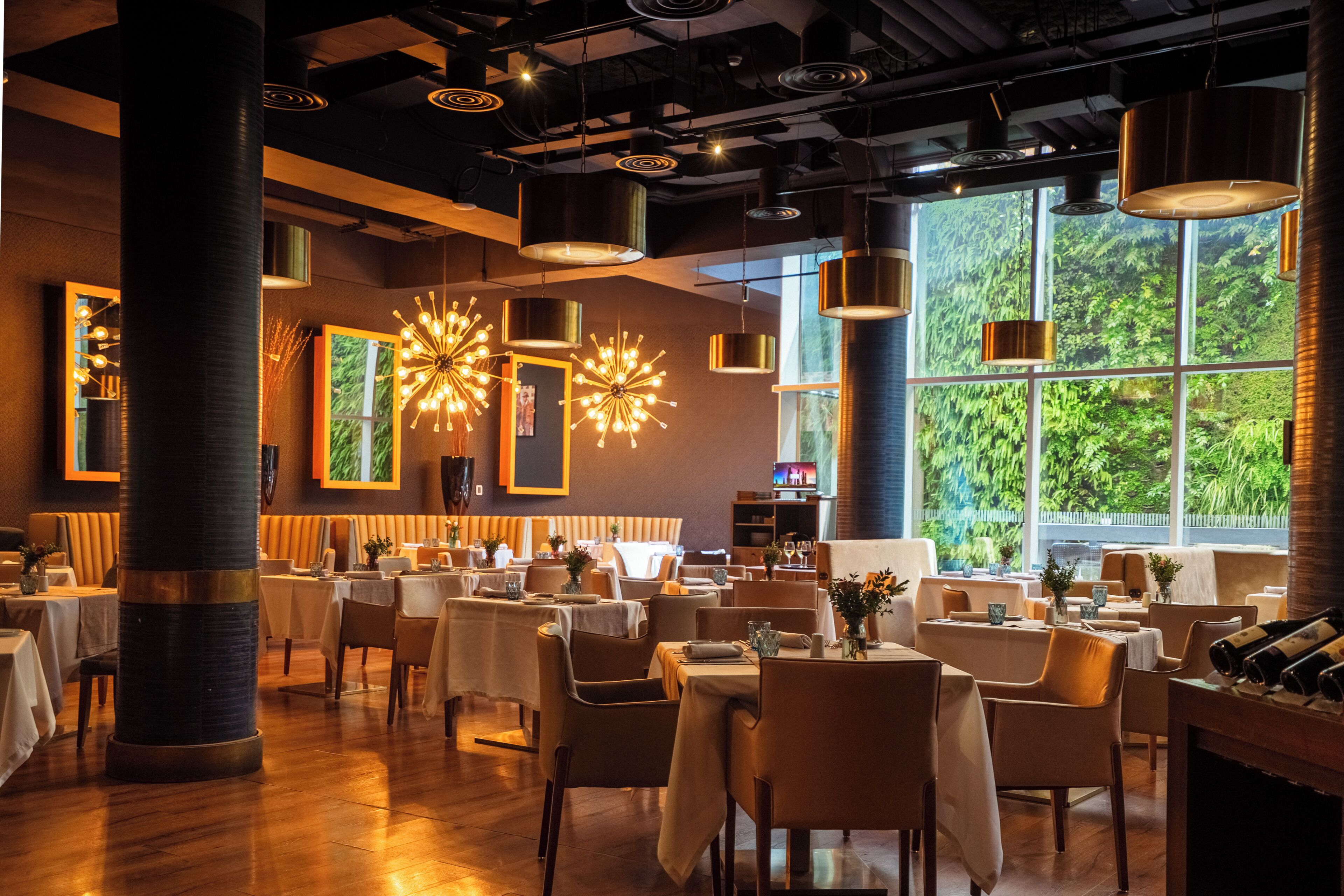 A restaurant interior with high ceilings, modern pendant lighting, and large windows offering a view of greenery. Tables are set with white tablecloths and chairs.