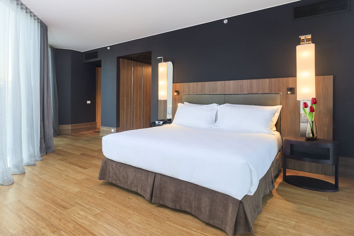 A modern hotel room with a large bed, wooden flooring, dark walls, and a nightstand with a vase of flowers.