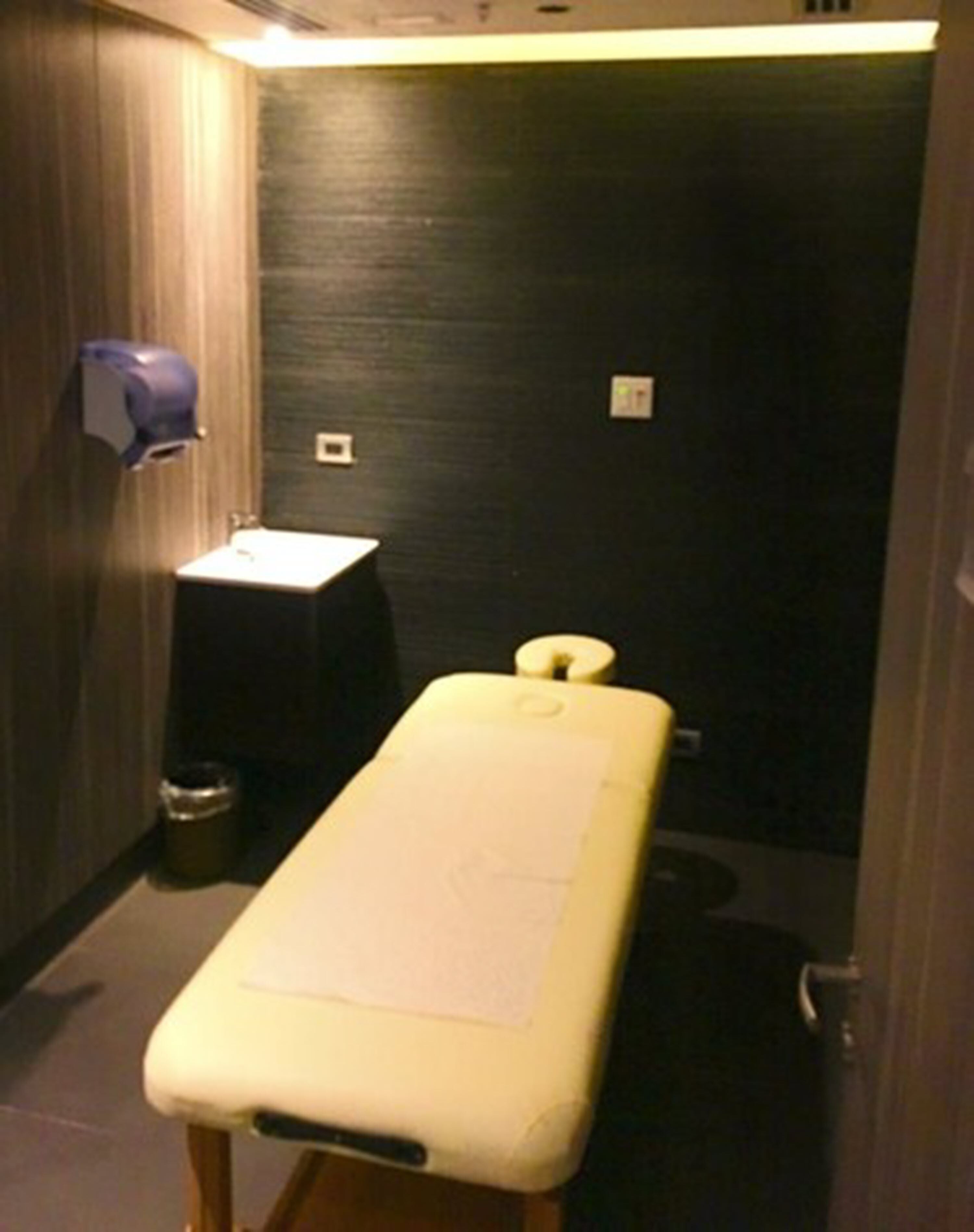 Una habitación pequeña y poco iluminada con una camilla, un lavabo y un cubo de basura.