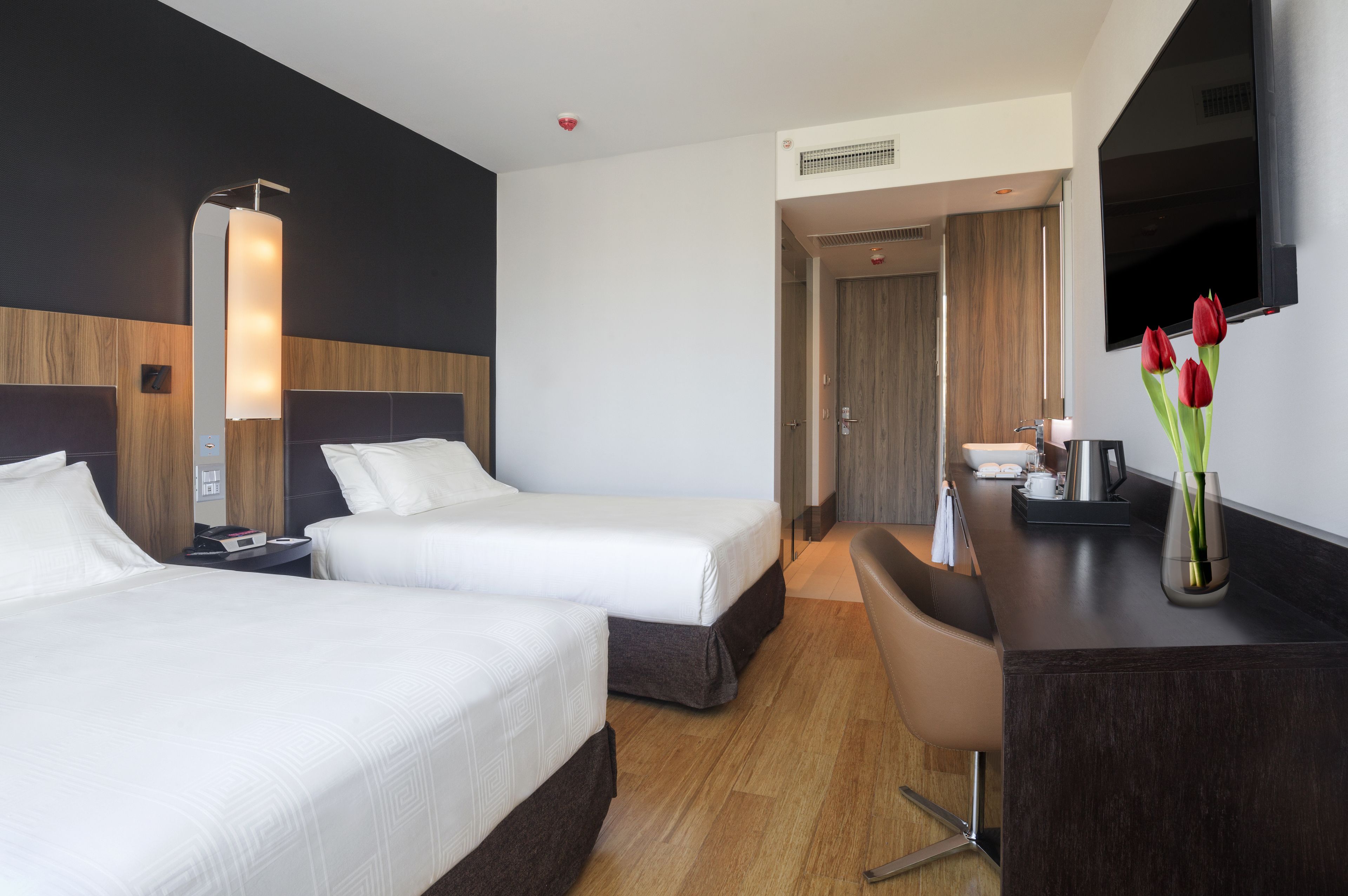 A hotel room with two beds, a desk, a chair, a TV, and a vase with flowers.