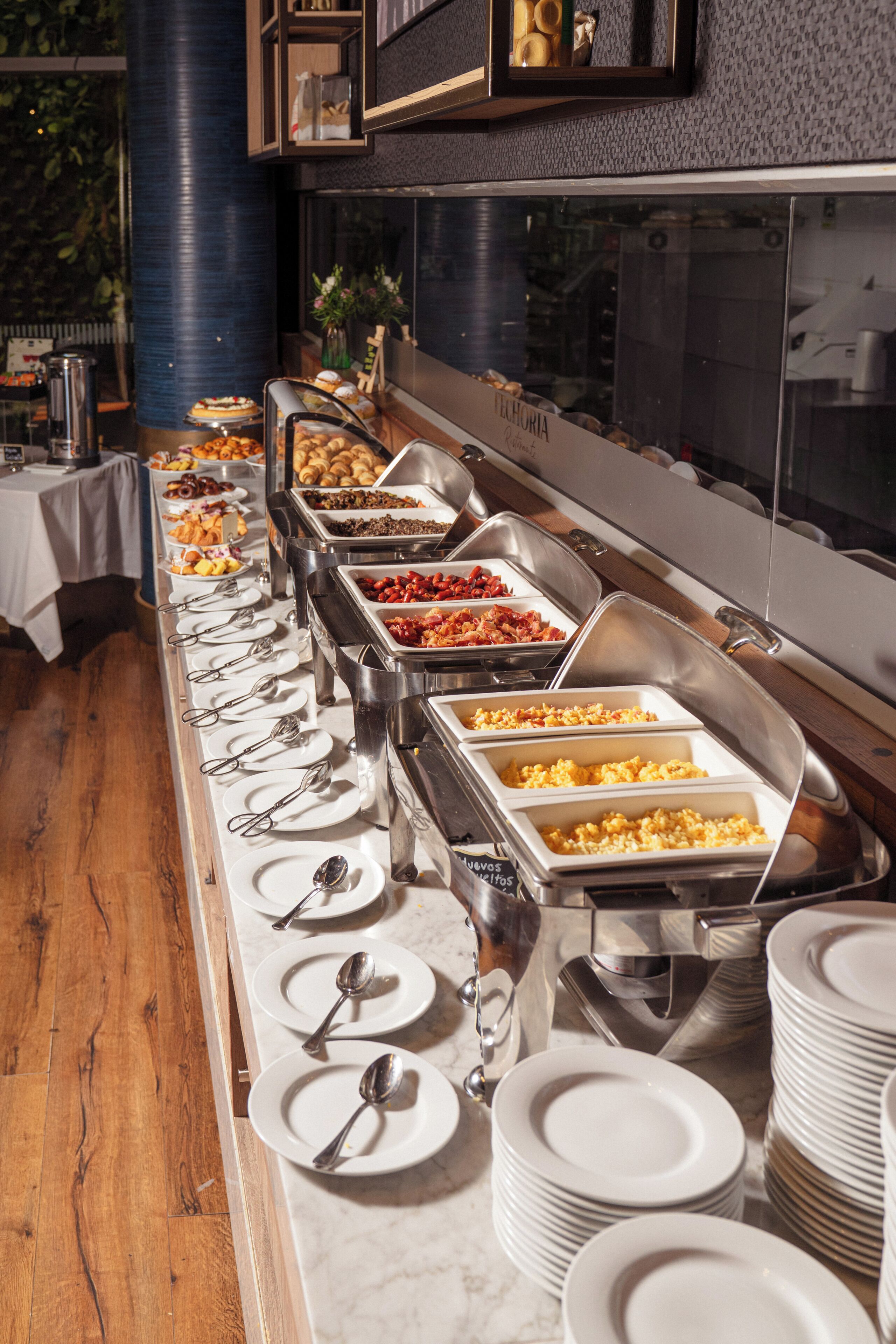A buffet setup with various food items in metal containers, arranged on a long table with plates and cutlery.