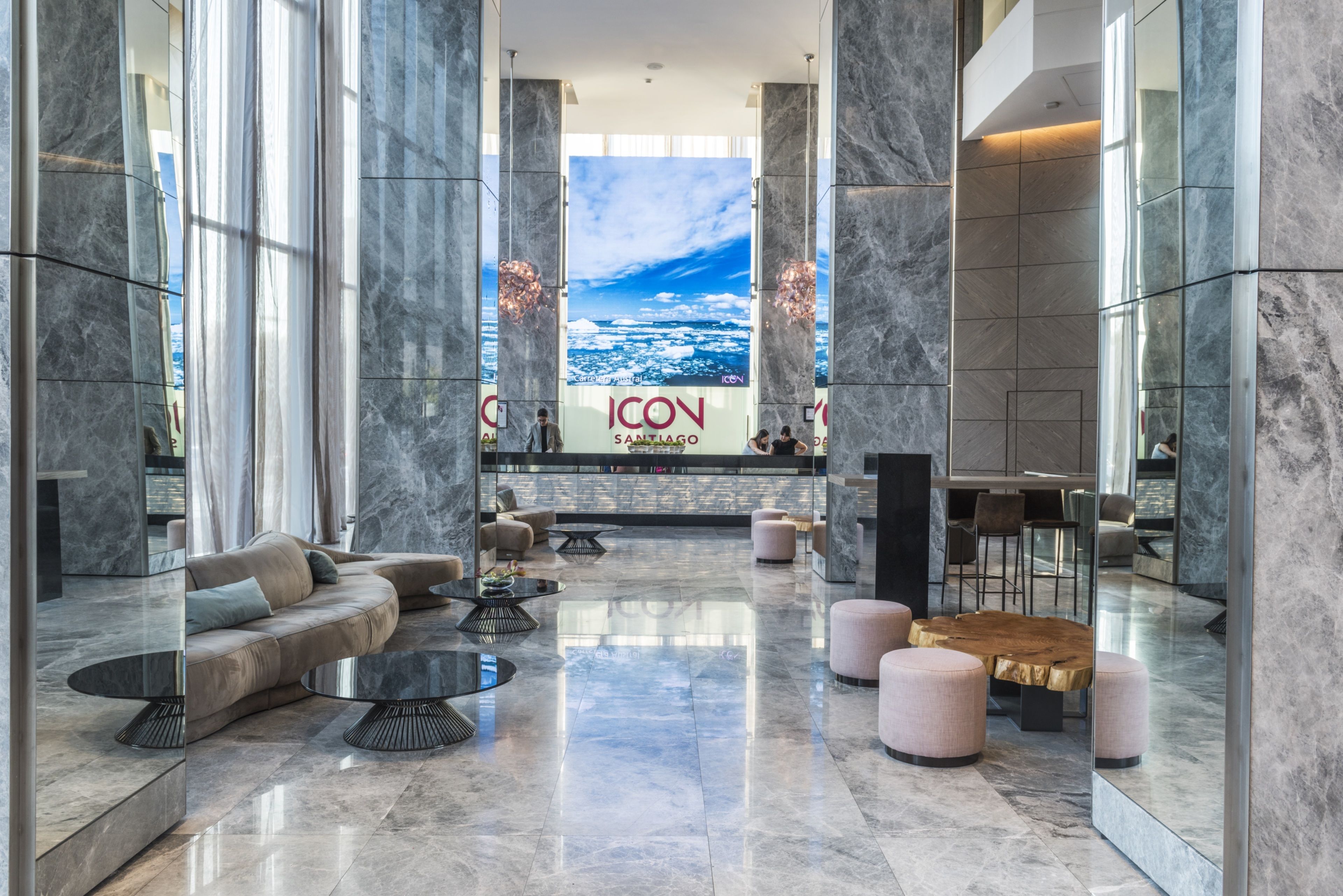 En rummelig lobby med marmorgulv, lædersofa, runde borde og et stort digitalt display, der viser 'ICON SANTIAGO'.