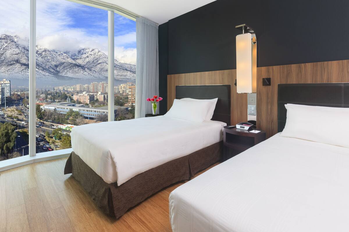 A hotel room with two beds, a large window with a city and mountain view, a nightstand, and a vase with flowers.
