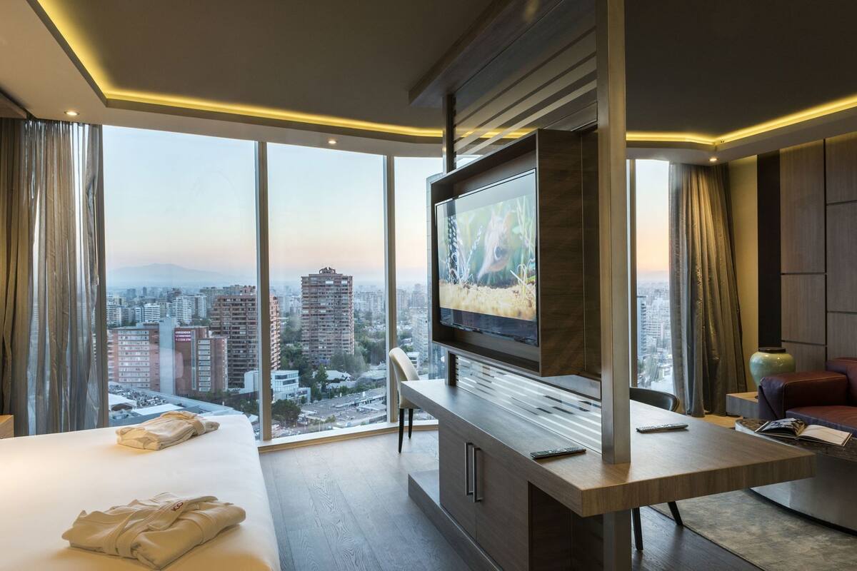 A modern hotel room with a large TV, a bed, a desk, and a view of the cityscape.
