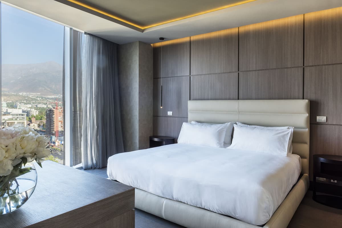 A modern hotel room with a large bed, a bedside table, a nightstand, and a view of a cityscape through large windows.