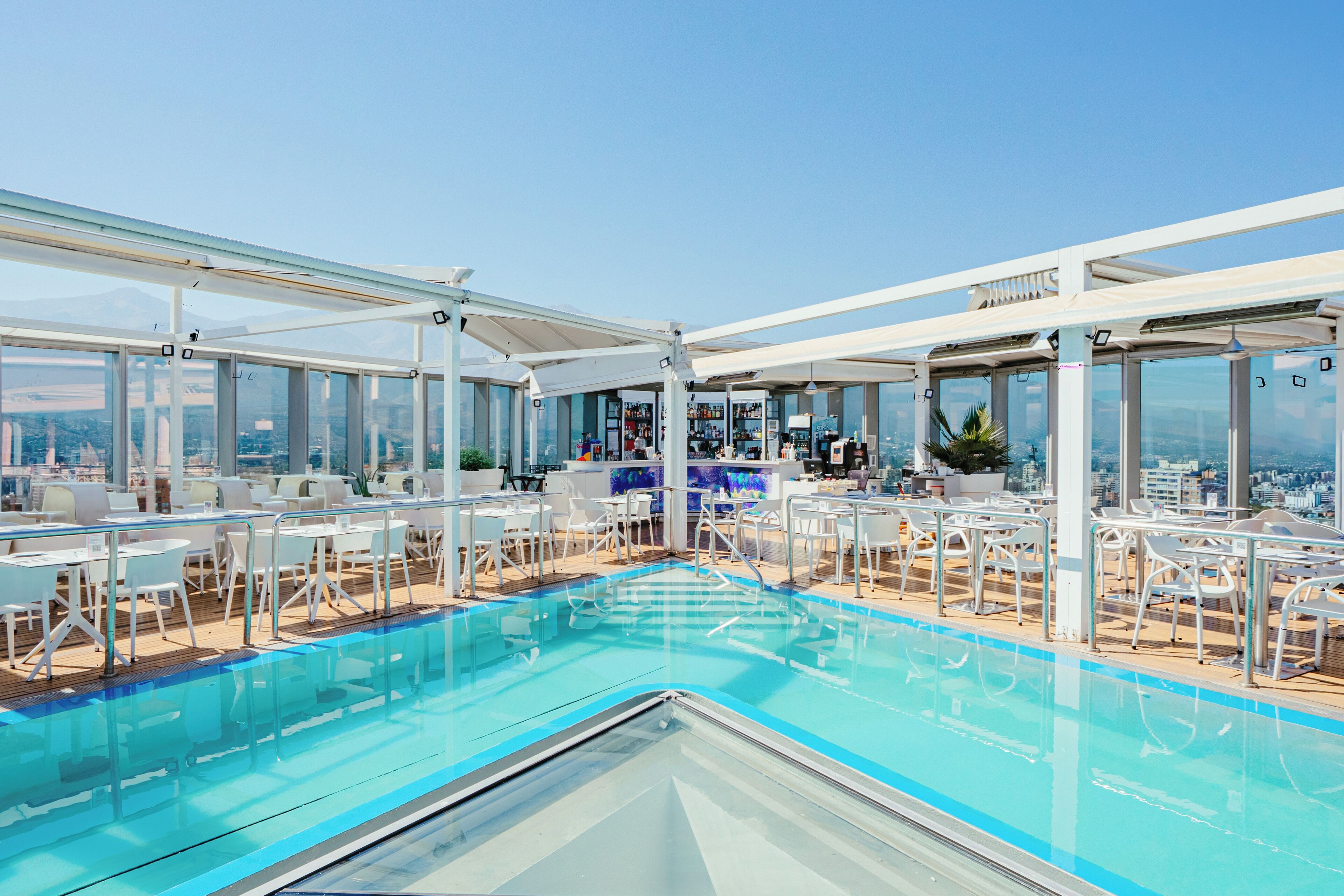 Un área de piscina en la azotea con asientos y un bar, con vistas al paisaje urbano.