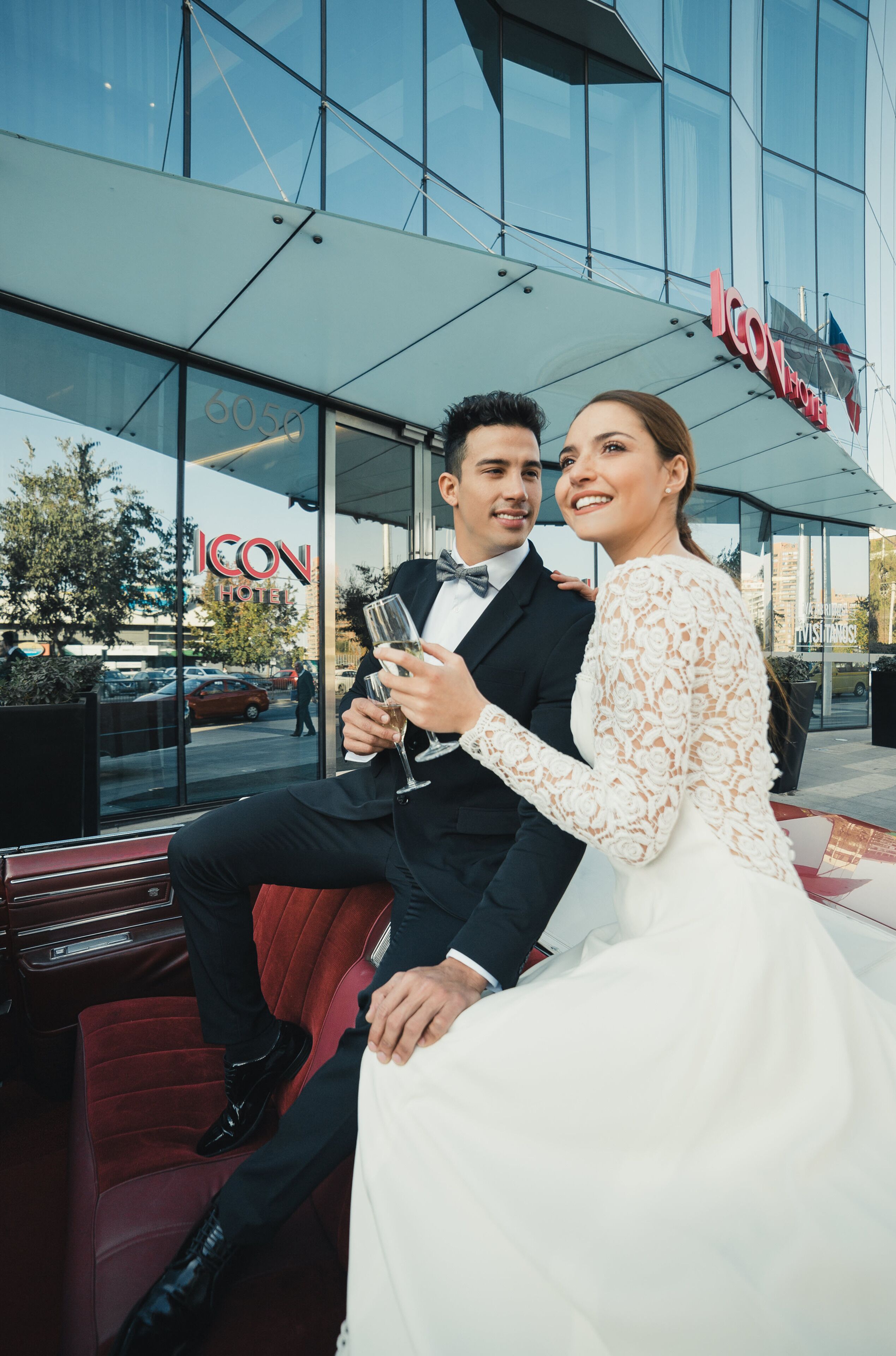 Una pareja sentada frente a un hotel de fachada de vidrio, con un letrero visible que dice "ICON HOTEL", y el reflejo de un coche en la ventana.
