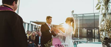 Una ceremonia de boda con una novia y un novio intercambiando alianzas, invitados sentados y un edificio moderno al fondo.