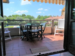 An outdoor patio with a table and chairs, a white chair, and a view of greenery and buildings in the background.