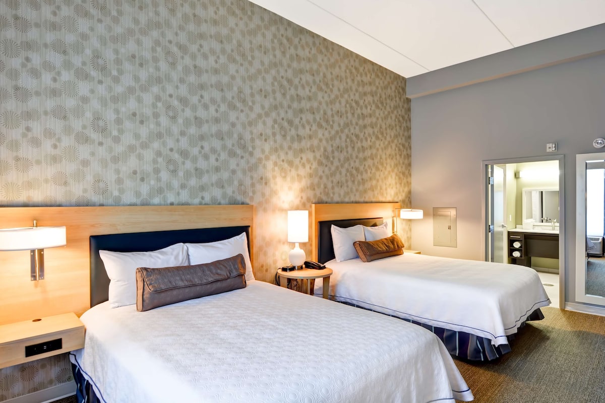 Une chambre d’hôtel avec deux lits, une table de chevet, une lampe et une salle de bain visible à travers une porte ouverte.