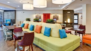 A modern hotel lobby with a green sofa, wooden tables, and chairs, and a red door in the background. - Home2 Suites by Hilton Stafford Quantico (Stafford)