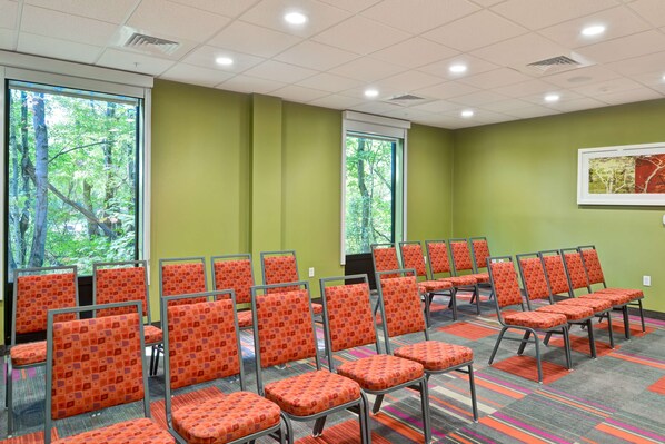 A conference room with rows of orange chairs, large windows with a view of trees, and a green wall.
