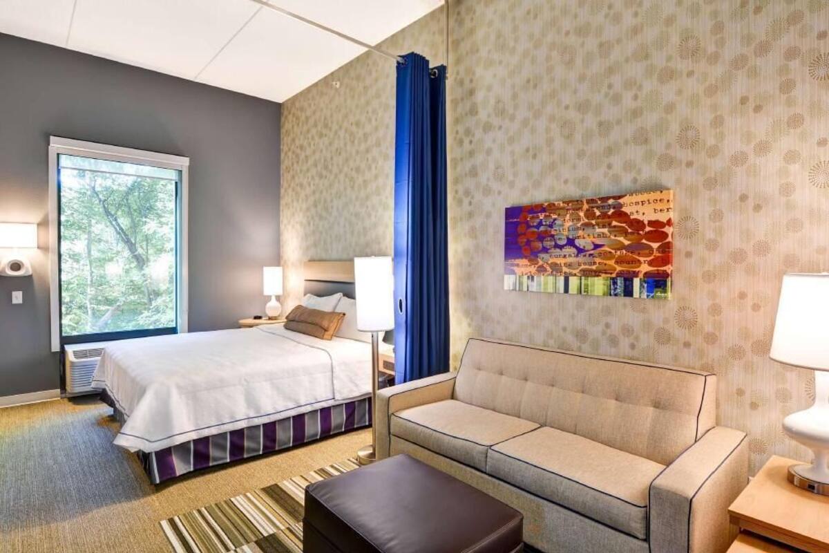 Une chambre d’hôtel comprenant un lit, un canapé, une petite table, une lampe, une fenêtre donnant sur des arbres et un mur orné d’une œuvre d’art colorée.