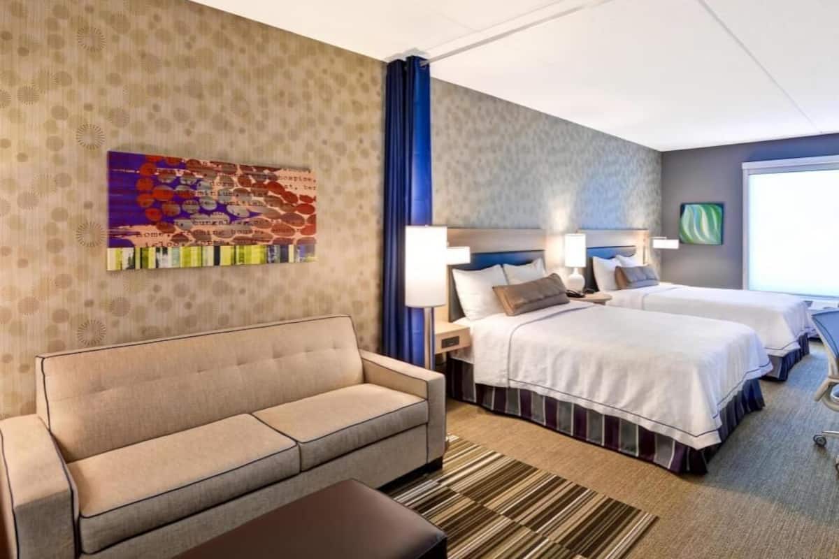 Une chambre d’hôtel avec un canapé, deux lits, une tête de lit, une lampe, une œuvre d’art murale et une fenêtre.