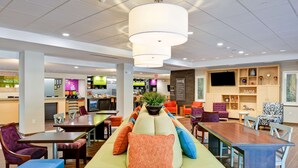 A modern hotel lobby with a green sofa, dining tables, and a TV mounted on the wall. - Home2 Suites by Hilton Stafford Quantico (Stafford)